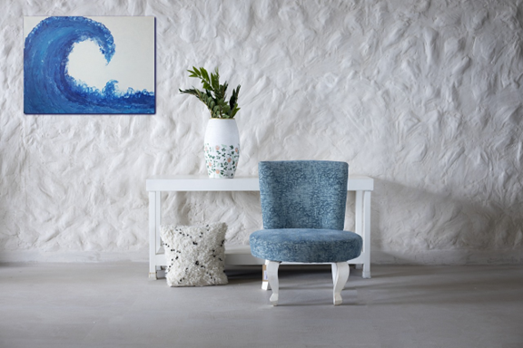 Une pièce composée d'un mur texturé blanc avec un cadre accroché en haut à gauche. Un meuble simple blanc se trouve à droite du cadre. Sur le meuble, il y a un pot blanc et bleu dans le bas avec une plante verte. Devant le meuble à droite, il y a une chaise bleue sans accoudoir. À gauche de la chaise, on y retrouve un coussin blanc avec des taches bleues dessus.