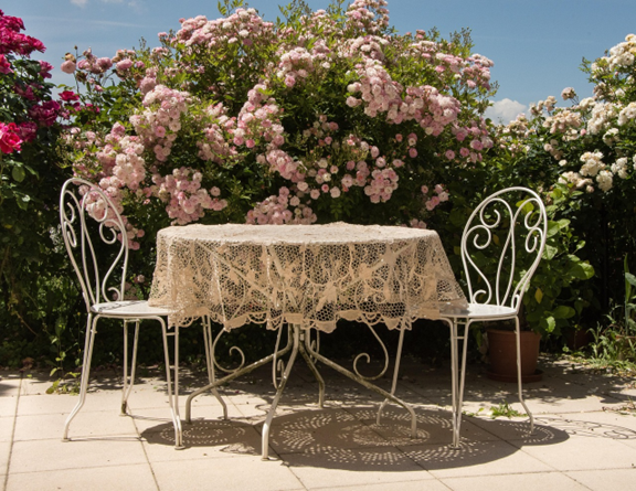 Il y a une table et deux chaises de style vintage blanche. Sur la table, il y a une nappe blanche avec des taches vertes. Le tout se trouve à l'extérieur devant un arbre à lilas fleuri de fleurs rose pâle.